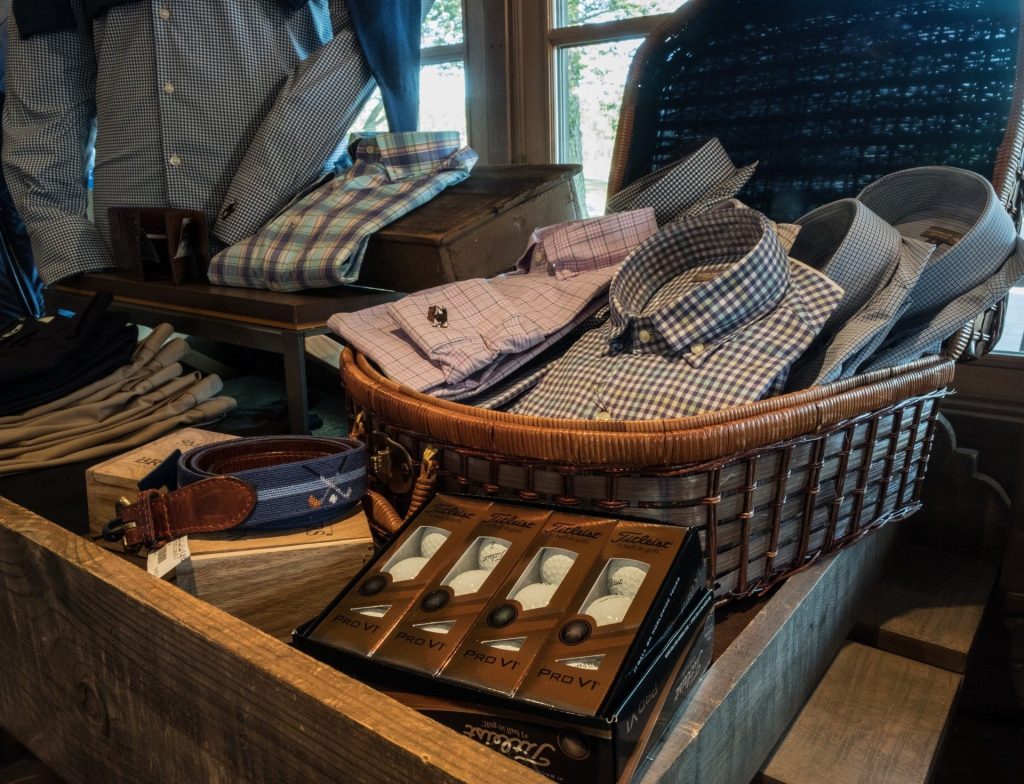 A wicker basket filled with neatly folded men's plaid button-down shirts, displayed alongside belts and Titleist Pro V1 golf balls in a rustic pro shop setting.